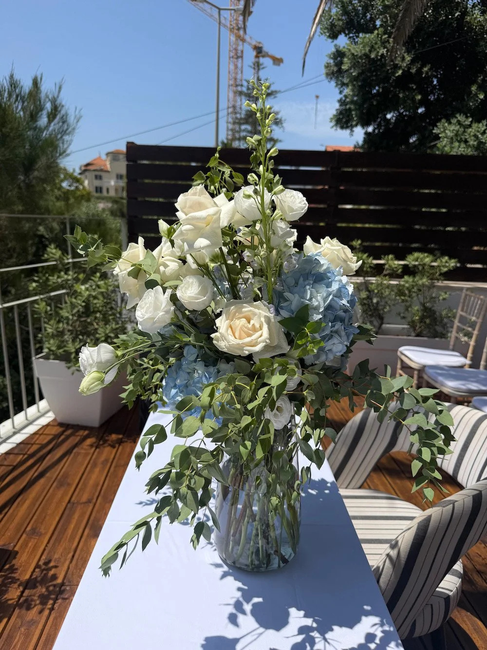 A glass vase with white roses, blue hydrangeas, and green foliage on a white table outdoors. In the background, patio furniture and a wooden privacy screen are visible, along with trees and a construction crane against a blue sky.