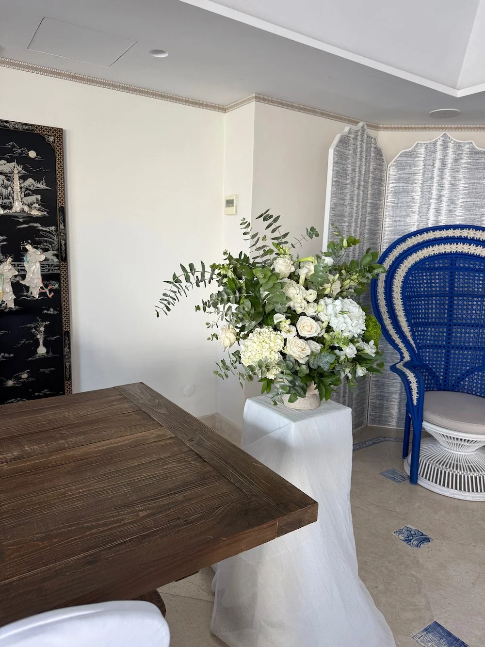 Interior room featuring a large floral arrangement of white roses and greenery on a white pedestal, with a wooden table in the foreground and decorative screens and a woven blue chair in the background.