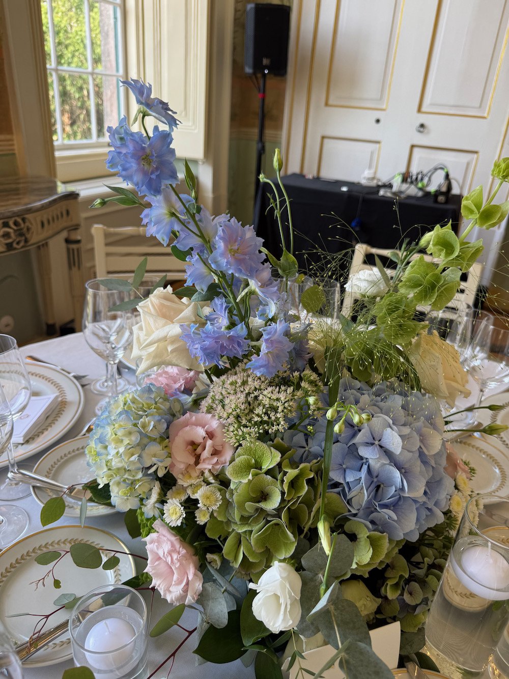 A floral centerpiece with blue, pink, green, and white flowers on a dining table inside a room with large windows, gold-trimmed walls, and a speaker in the background.