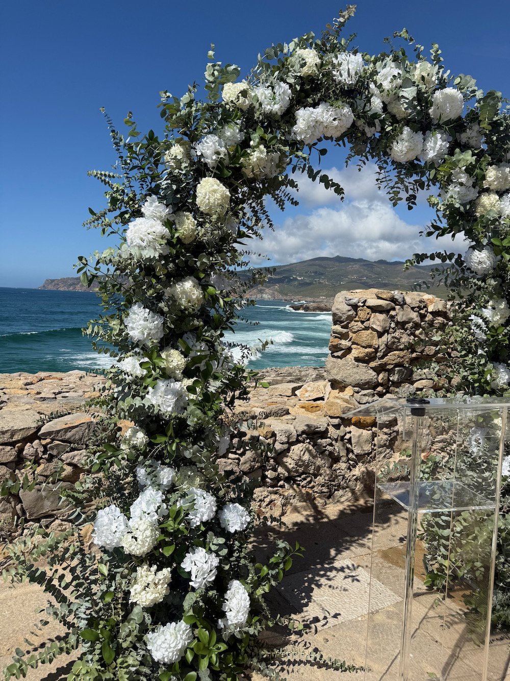 A floral arch made of white flowers and green leaves set against a beach with rocky shoreline, ocean waves, and a mountain in the background under a blue sky with some clouds.