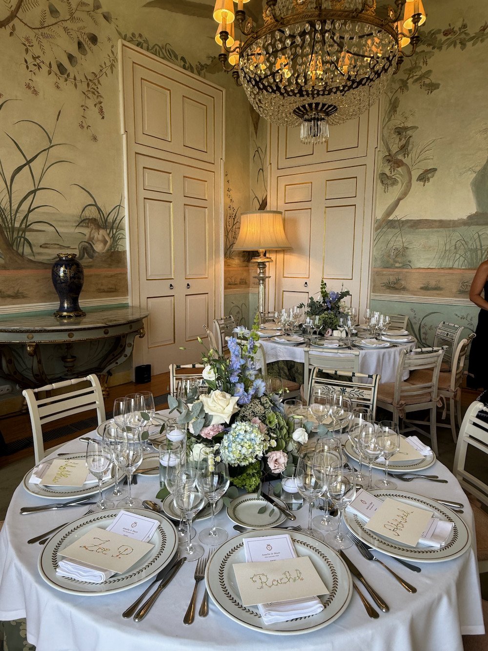 Elegant dining room with round table set for a formal event, decorated with floral centerpieces, multiple wine glasses, and place cards, surrounded by classic chairs, with a chandelier hanging from the ceiling and muraled walls.