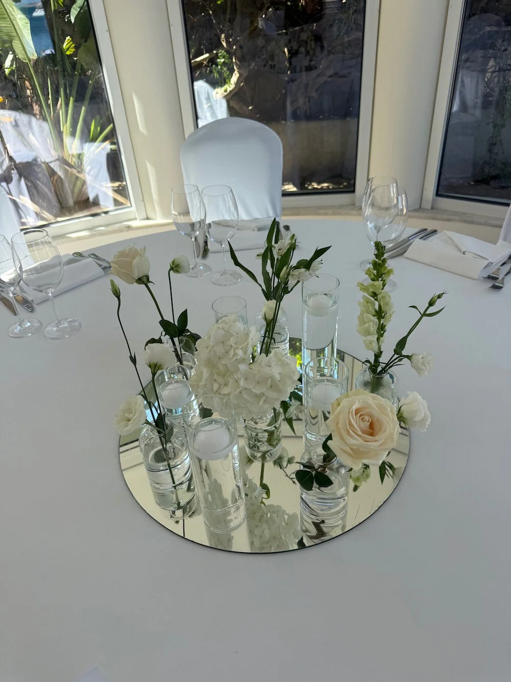Elegant round table set with white tablecloth, floral centerpiece with white roses and hydrangeas, surrounded by glassware and silverware, location by large windows with outdoor greenery.