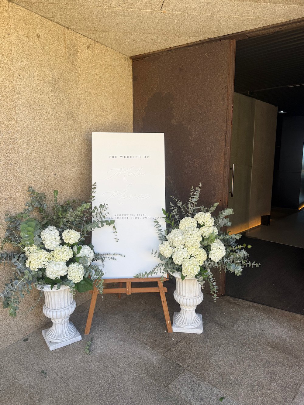 Wedding sign on an easel with white floral arrangements in white urns at each side placed outside an entrance