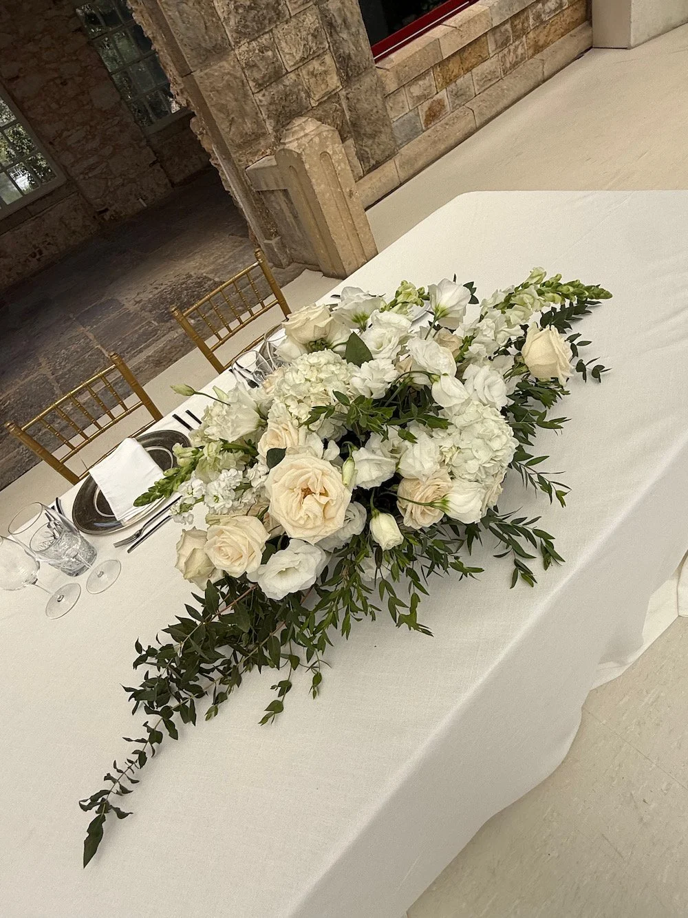 A long floral centerpiece with white and cream-colored flowers and green foliage on a white tablecloth, set for a formal event.
