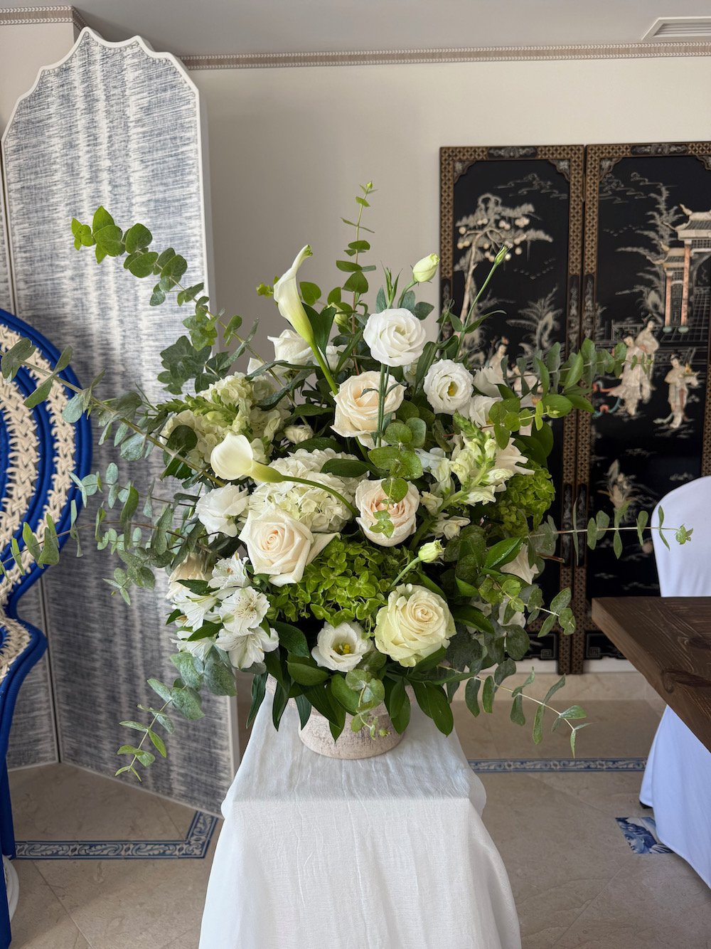 A large floral arrangement with white roses, lilies, and green foliage in a beige vase on a white cloth-covered table.