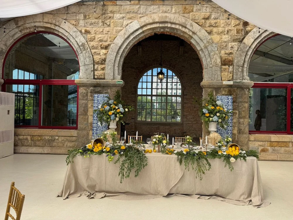 Table decorated with yellow and white floral arrangements, lemons, and lit candles, set in a rustic brick building with arched windows and a table with a beige tablecloth.