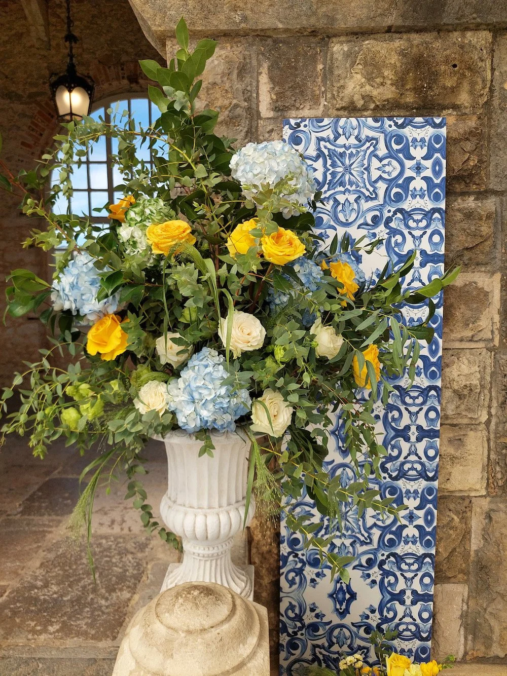 A large floral arrangement with blue hydrangeas, yellow roses, and white roses in a tall white vase, placed beside a decorative blue and white patterned panel against a stone wall background.