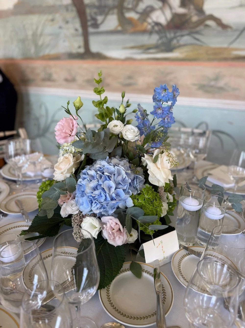 Centerpiece of a floral arrangement with blue hydrangeas, white roses, pink lisianthus, blue delphiniums, and greenery on a dining table set with plates, silverware, wine glasses, and votive candles in a decorated dining room.