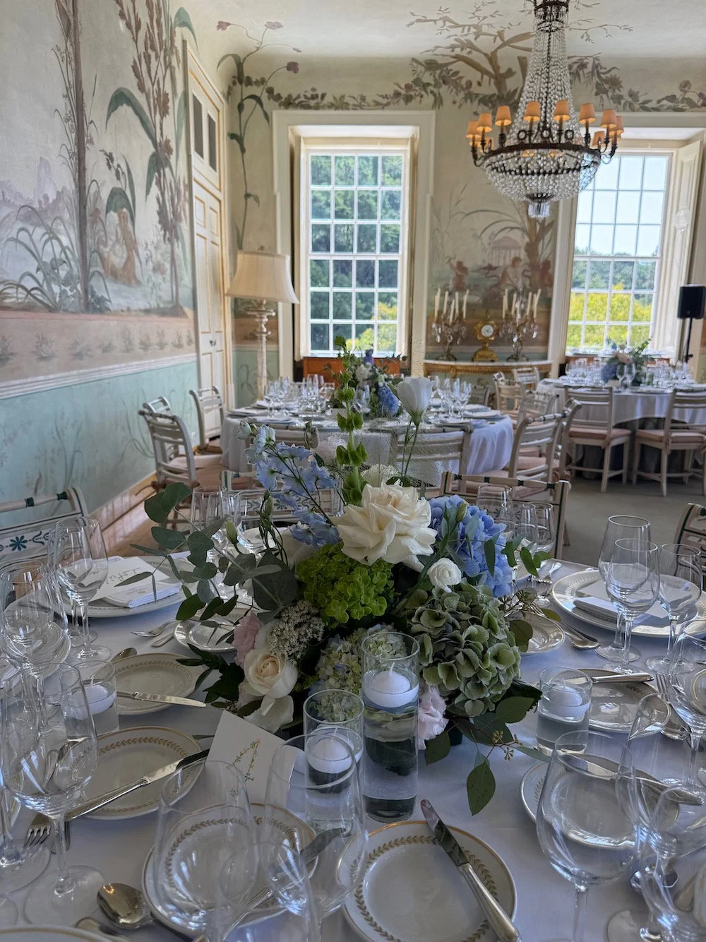 Elegant banquet room with a chandelier, floral wall murals, and a table set with glassware, silverware, and a centerpiece of white, blue, and green flowers.