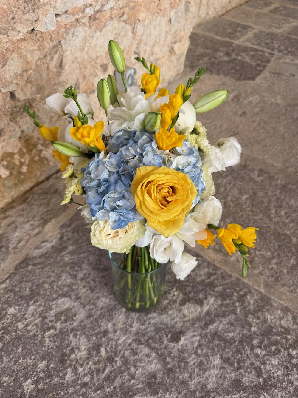 A bouquet of mixed flowers including yellow roses, white lisianthus, blue hydrangeas, and yellow freesias in a clear glass vase on a stone surface with a brick wall background.