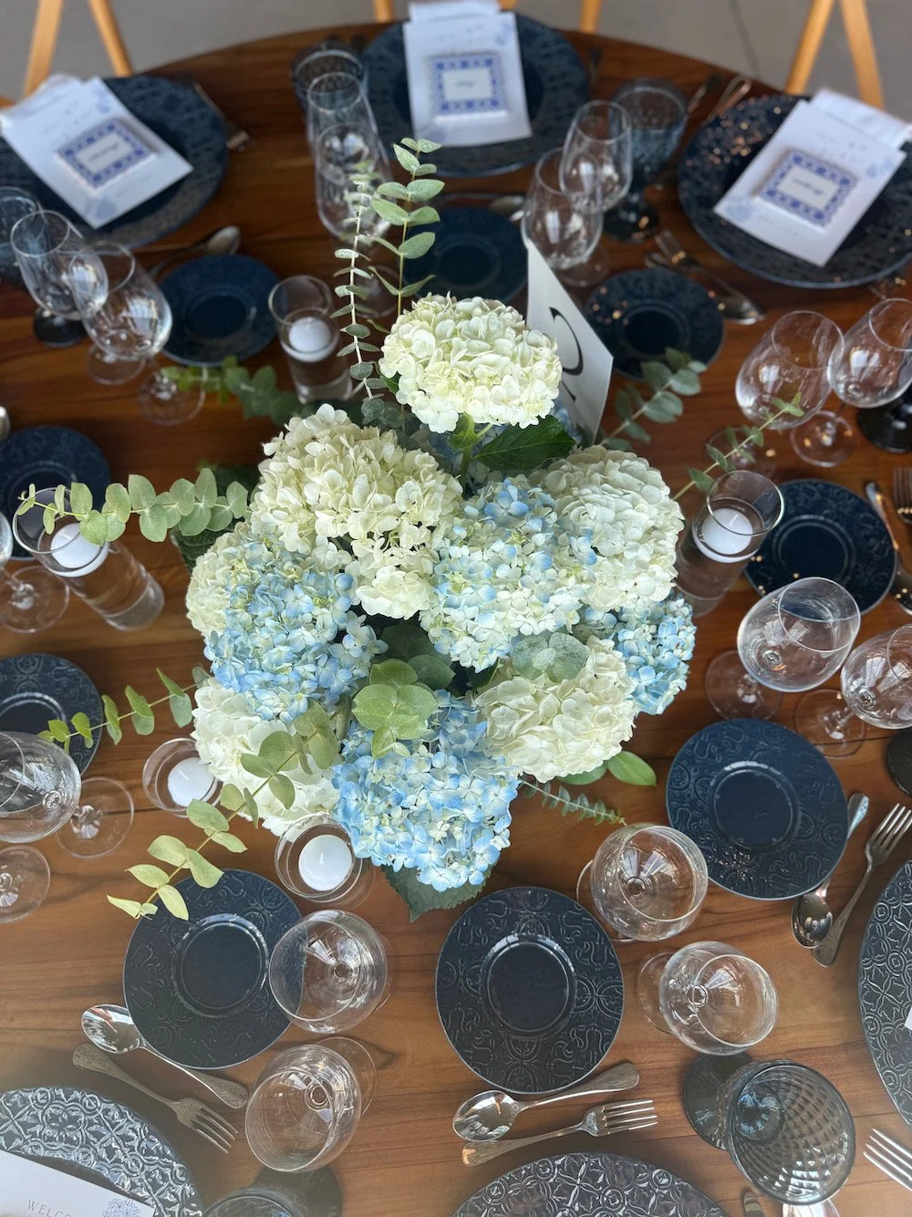 Elegant dinner table setup with a large centerpiece of white and blue hydrangeas, surrounded by black plates, clear glasses, and silverware on a wooden table.