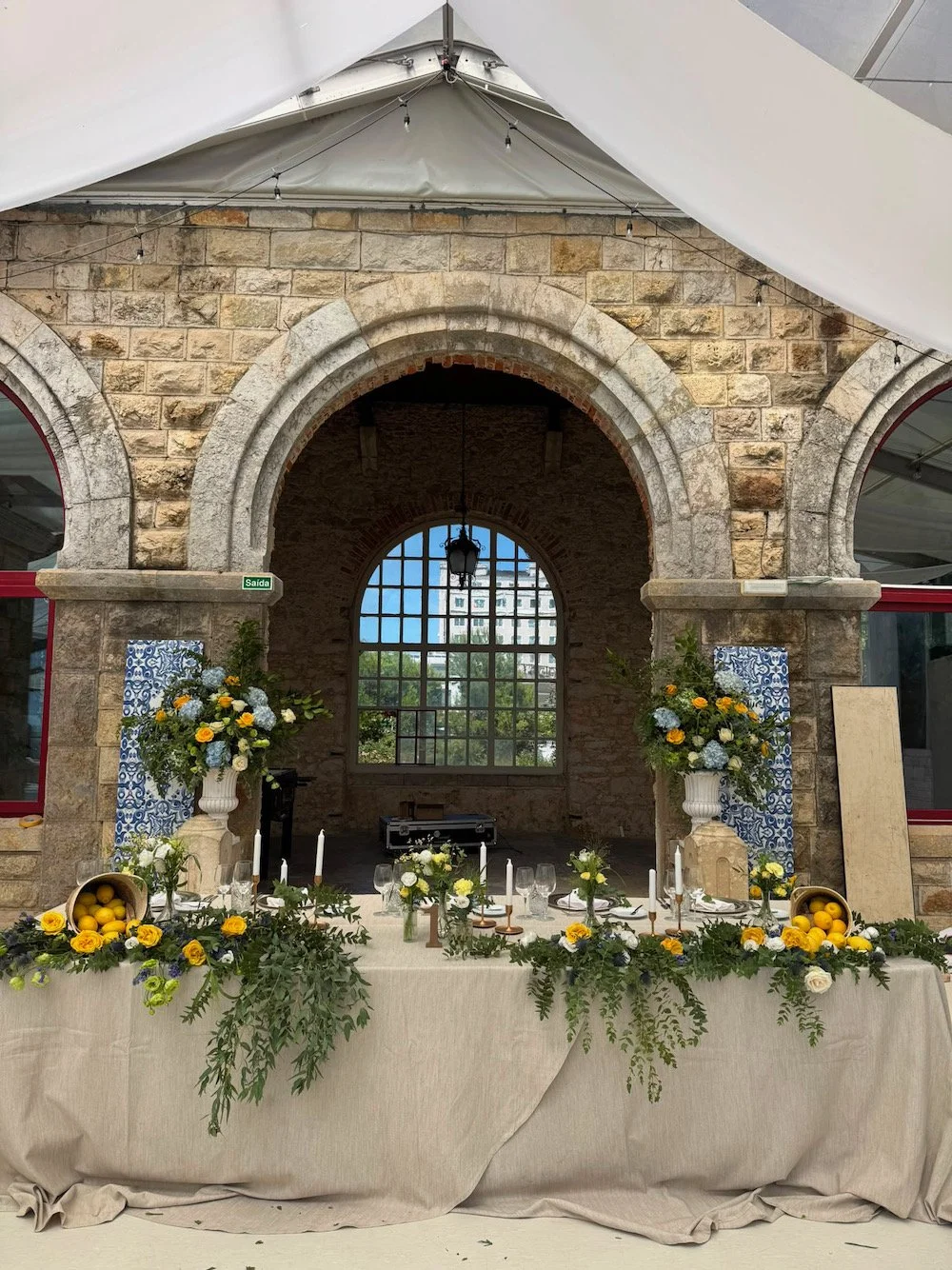 Decorated banquet table with floral arrangements and lemons, inside a rustic stone building with arches and large windows.