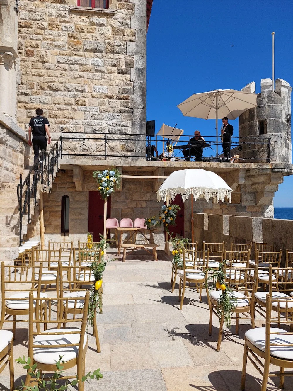 An outdoor wedding setup on a stone terrace with chairs decorated with greenery and lemons, a white parasol, and a table with flowers. There's a stone building in the background with musicians on a balcony and staff preparing for the event.
