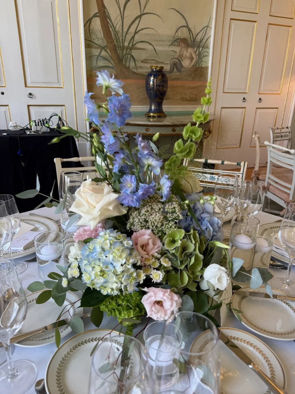 Elegant dining table with a floral centerpiece including white roses, light purple flowers, and greenery, set with gold-trimmed china and multiple wine glasses in a refined room with wall panels and a decorative background.