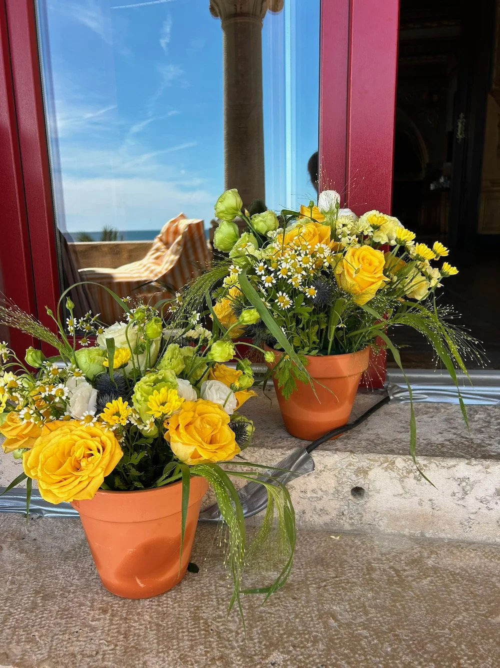 Two terracotta pots with yellow and white flowers, resting on a stone surface near a red-framed window. Through the window, a sunny sky, balcony with striped fabric, and a column are visible.