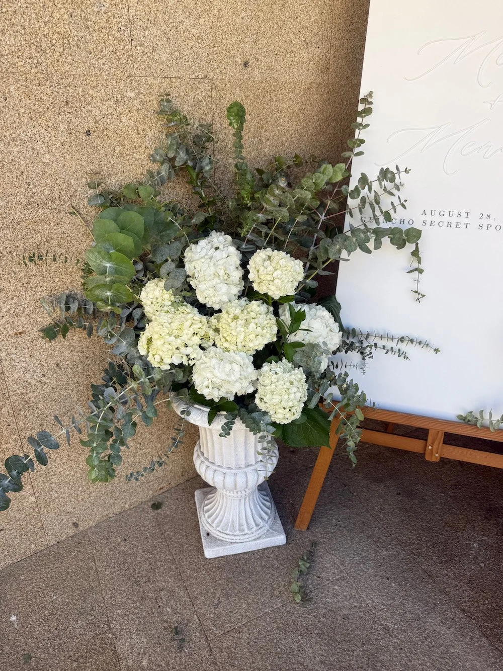 Arrangement of white hydrangea flowers and green foliage in a white, classical-style urn on a gray stone floor.