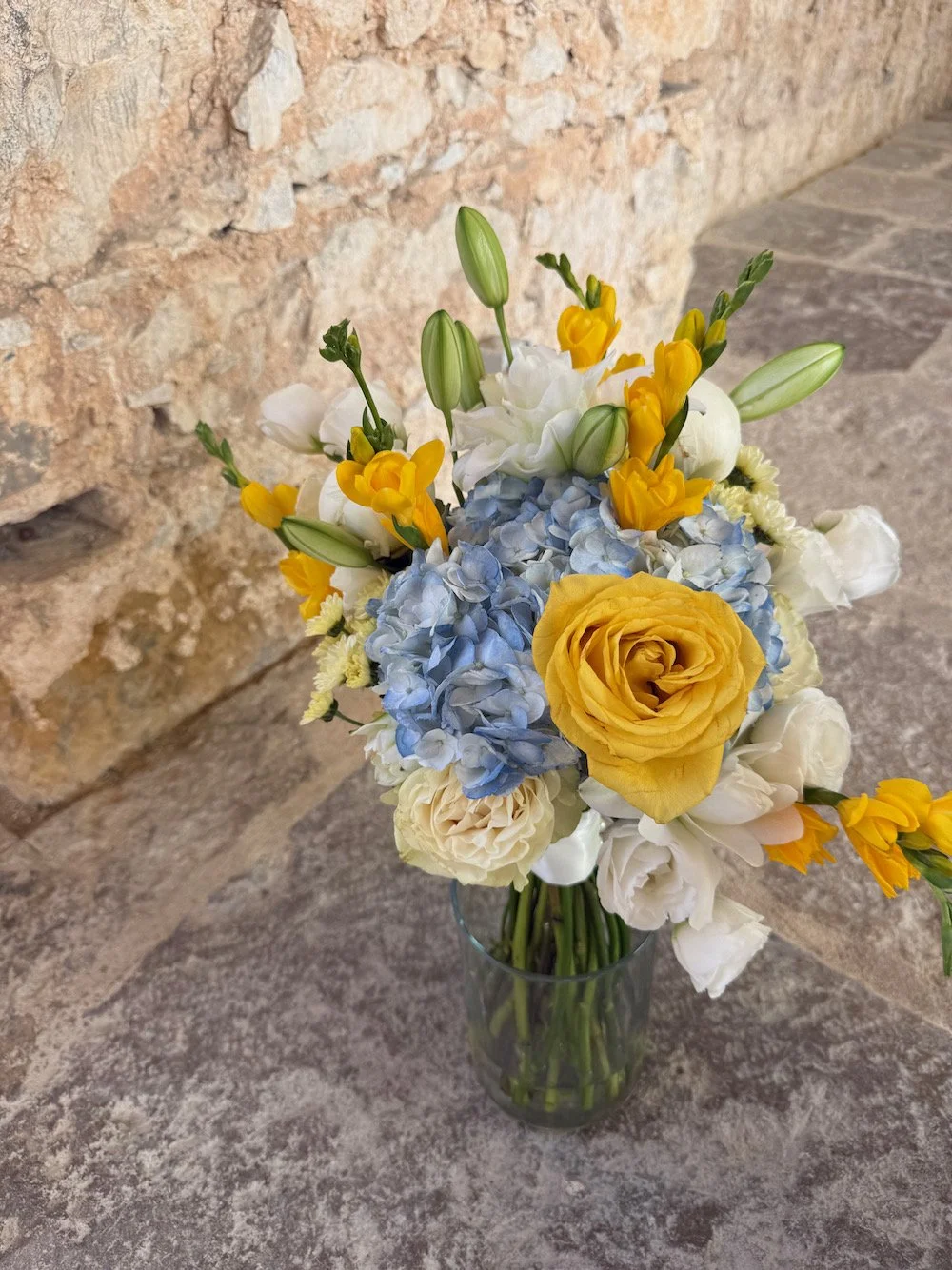A bouquet of flowers in a glass vase, featuring yellow roses, blue hydrangeas, white carnations, and yellow freesias, placed on a stone surface against a rustic stone wall.