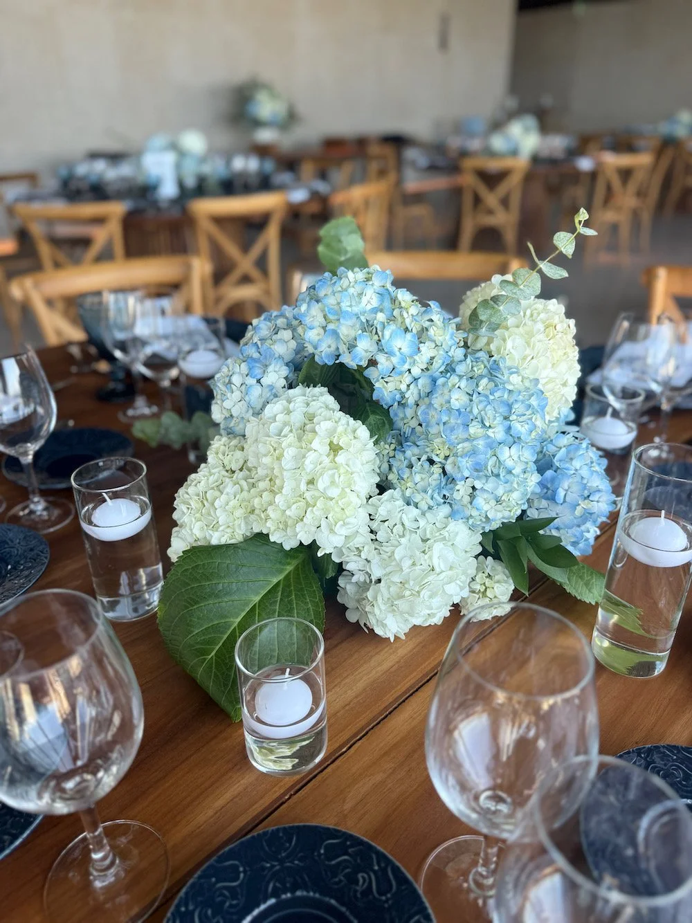 Elegant tablesetting with a floral centerpiece of white and light blue hydrangeas, surrounded by wine glasses, water glasses with floating candles, dark blue plates, and wooden chairs in a rustic indoor setting.