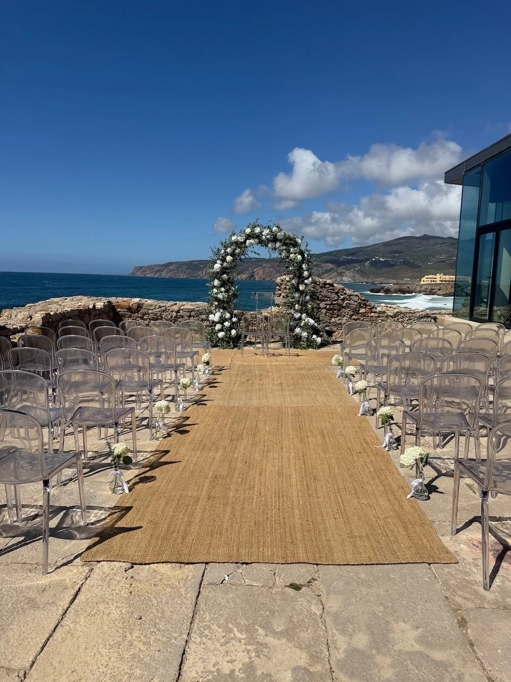Outdoor wedding setup with rows of transparent chairs on either side of a beige carpet, leading to a floral arch on a rocky coastline with ocean waves and mountains in the background under a partly cloudy sky.