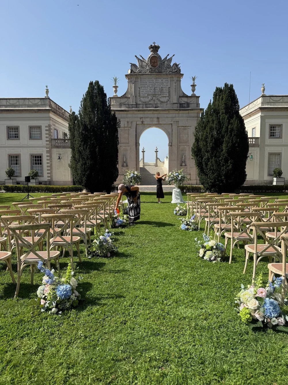 Setup for outdoor wedding ceremony on a lush green lawn with chairs arranged on either side of a flower-covered aisle leading to a historical stone archway with decorative elements, flanked by two large trees, under a clear blue sky.