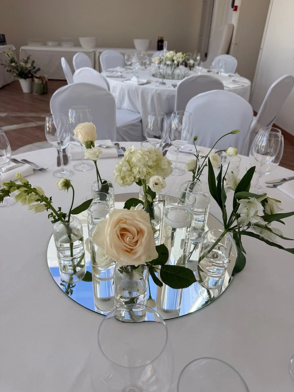 Elegant banquet table decorated with white roses and other white flowers in glass vases, set for a formal event with white tablecloths, glassware, and silverware.