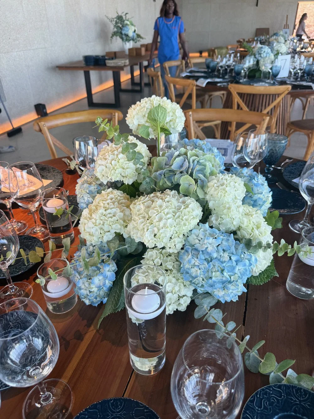A wedding or event table decorated with a centerpiece of white and light blue hydrangeas, surrounded by glassware, candles, and place settings on a wooden table, with chairs and another table in the background.