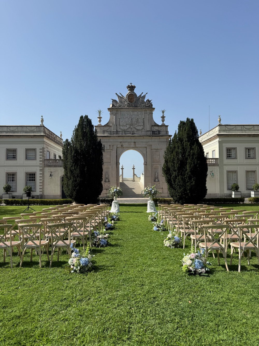 An outdoor wedding setup with rows of chairs and floral arrangements on a grassy lawn, leading to an ornate archway with classical architecture and sculptures, under a clear blue sky.