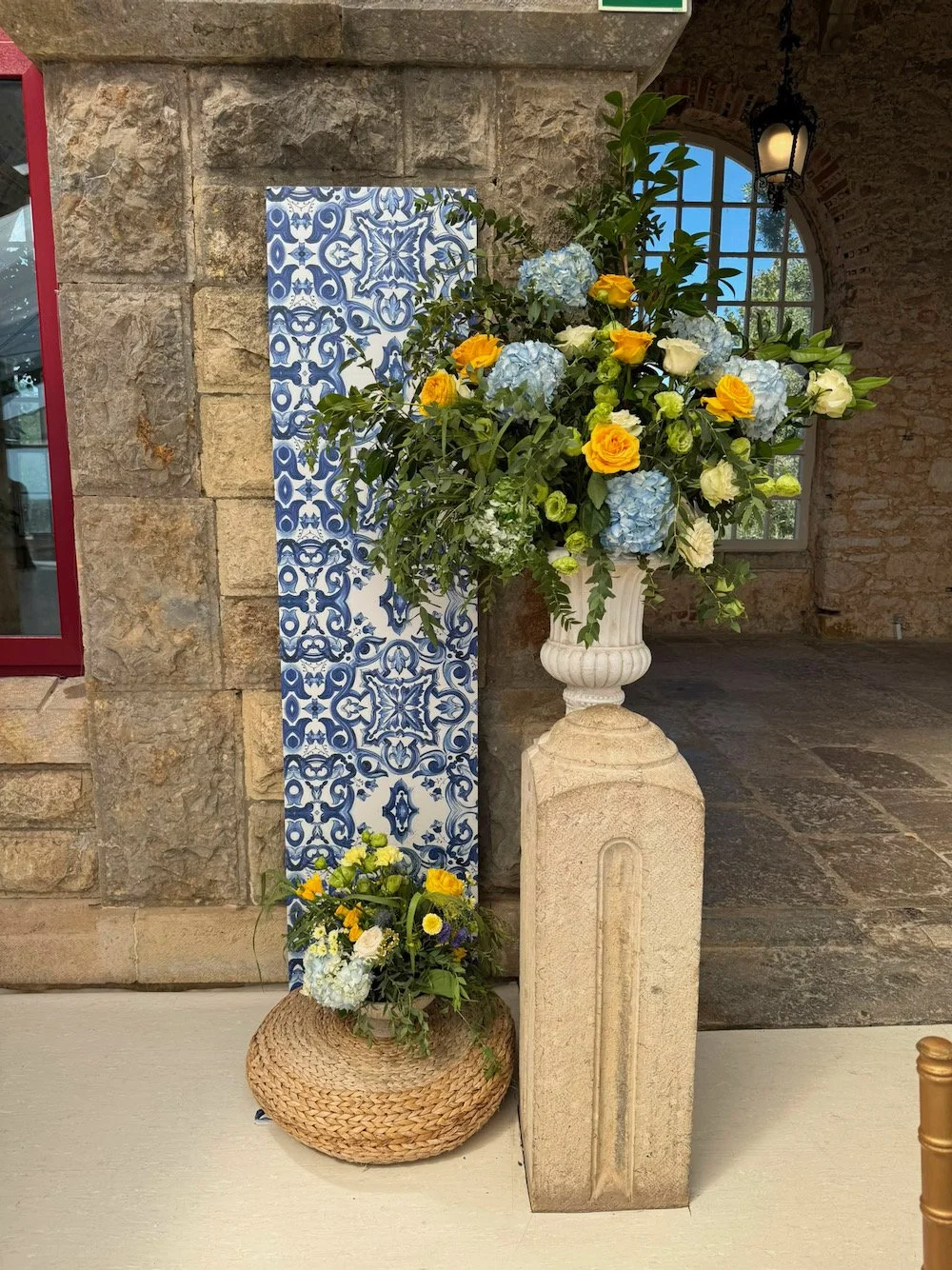 Large floral arrangement with blue hydrangeas, yellow roses, and white flowers in a white vase on a stone pedestal. Smaller flower arrangement at base on woven pouf, both placed near a decorative blue and white ceramic tile against a stone wall inter