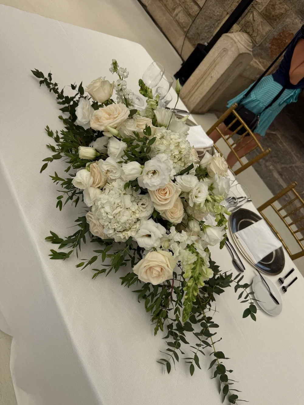 A white floral arrangement with roses, hydrangeas, and greenery on a white table setting at a formal event.