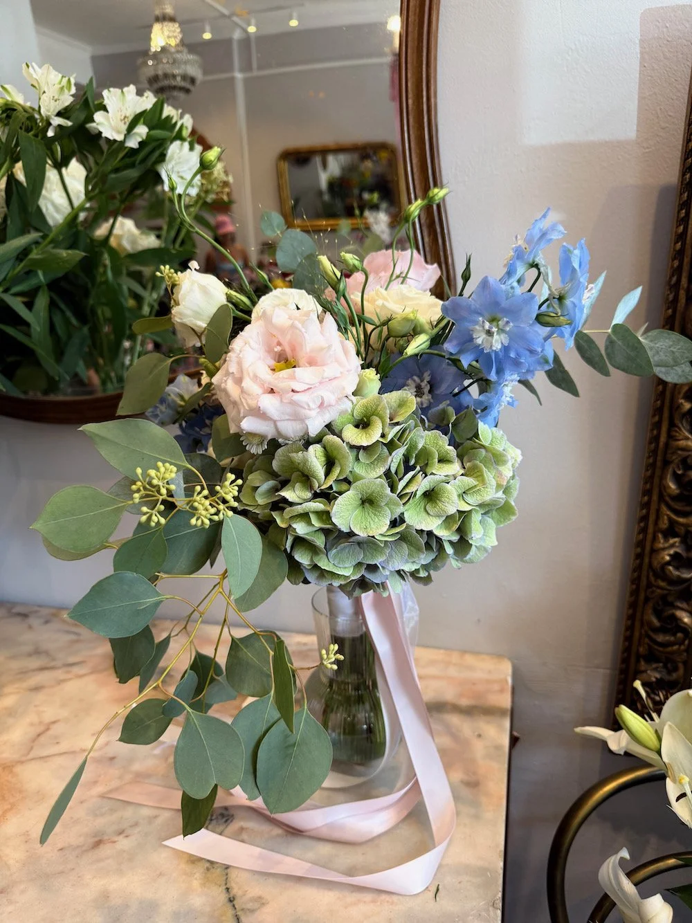 A bouquet of mixed flowers including white lisianthus, pink lisianthus, light blue delphiniums, and green hydrangeas with green foliage in a clear glass vase, tied with a light pink ribbon.