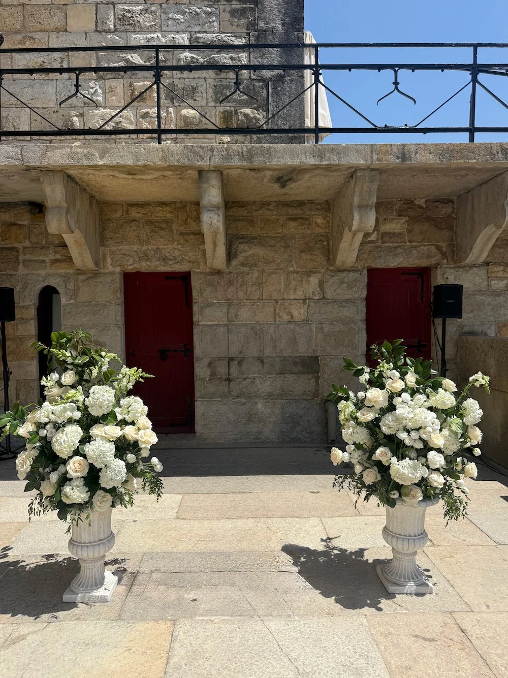 Two white flower arrangements in vases placed outside a stone building with two red doors for a wedding or special event.