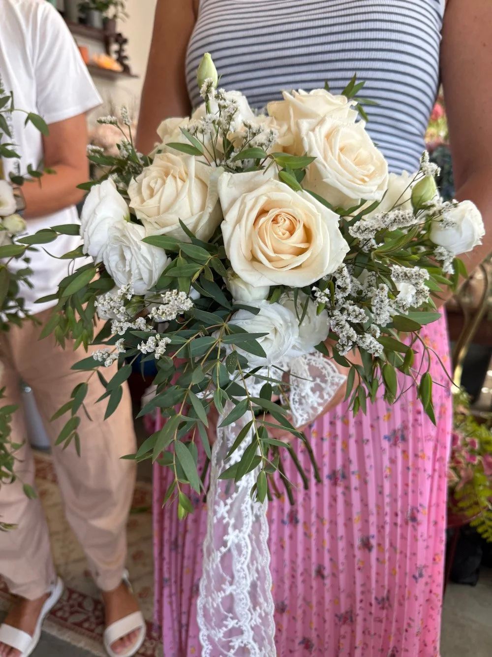 Person holding a bouquet of white roses, greenery, and white accent flowers, with lace ribbon, in a casual setting.