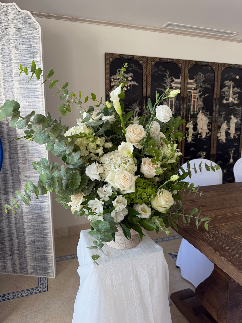 A large bouquet of white roses, lilies, hydrangeas, and greenery arranged in a vase on a white cloth-covered stand in a room with Asian-inspired artwork on a folding screen in the background.