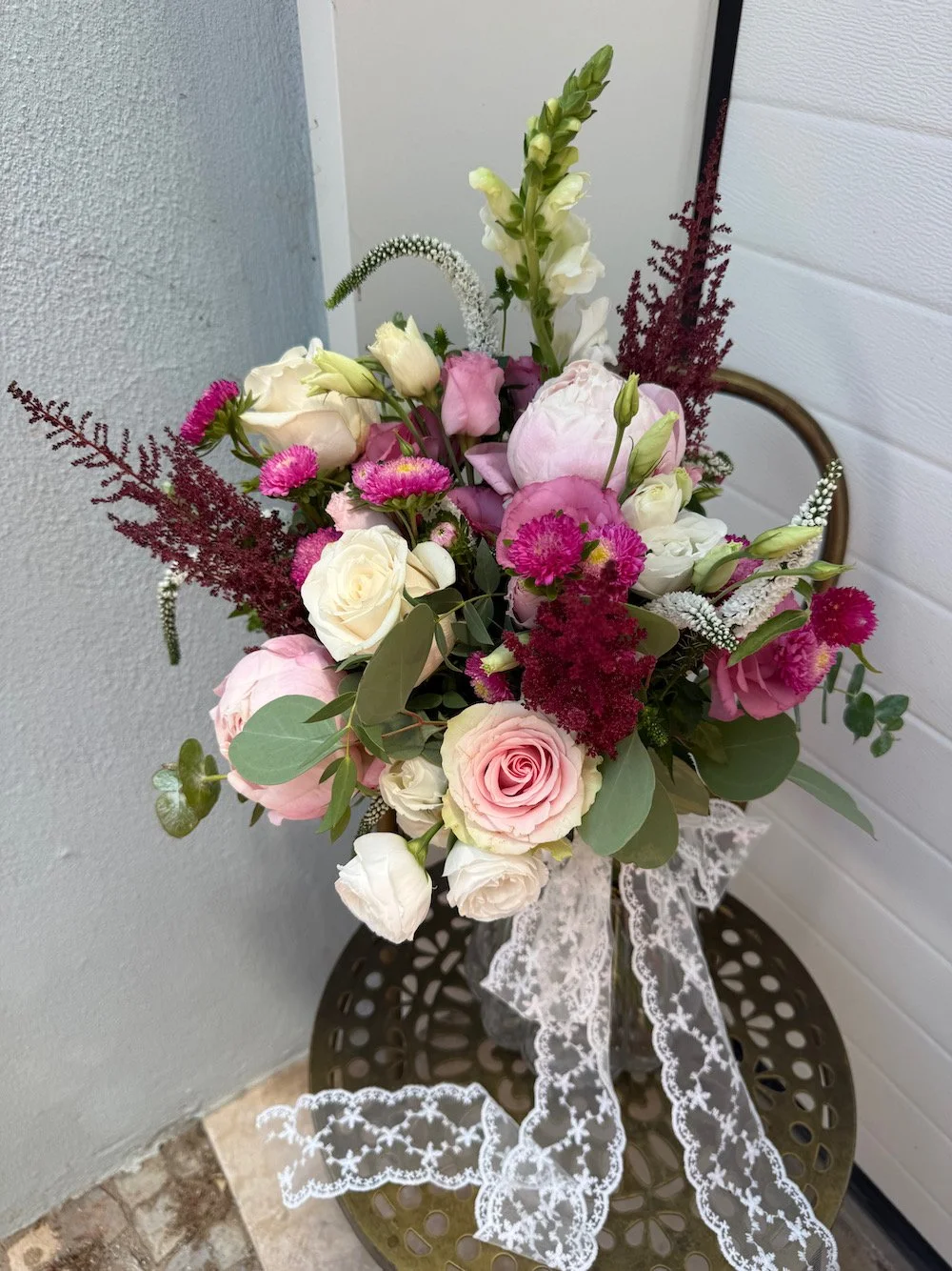 Bouquet of pink and white roses, white lisianthus, and dark red astilbe arranged with green foliage, tied with a white lace ribbon on a round metal table.