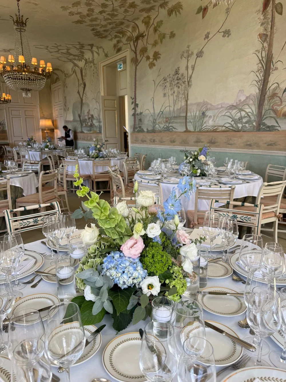 Elegant banquet table setup with floral centerpiece, fine china, and multiple glasses in a decorated room with mural walls and chandeliers.