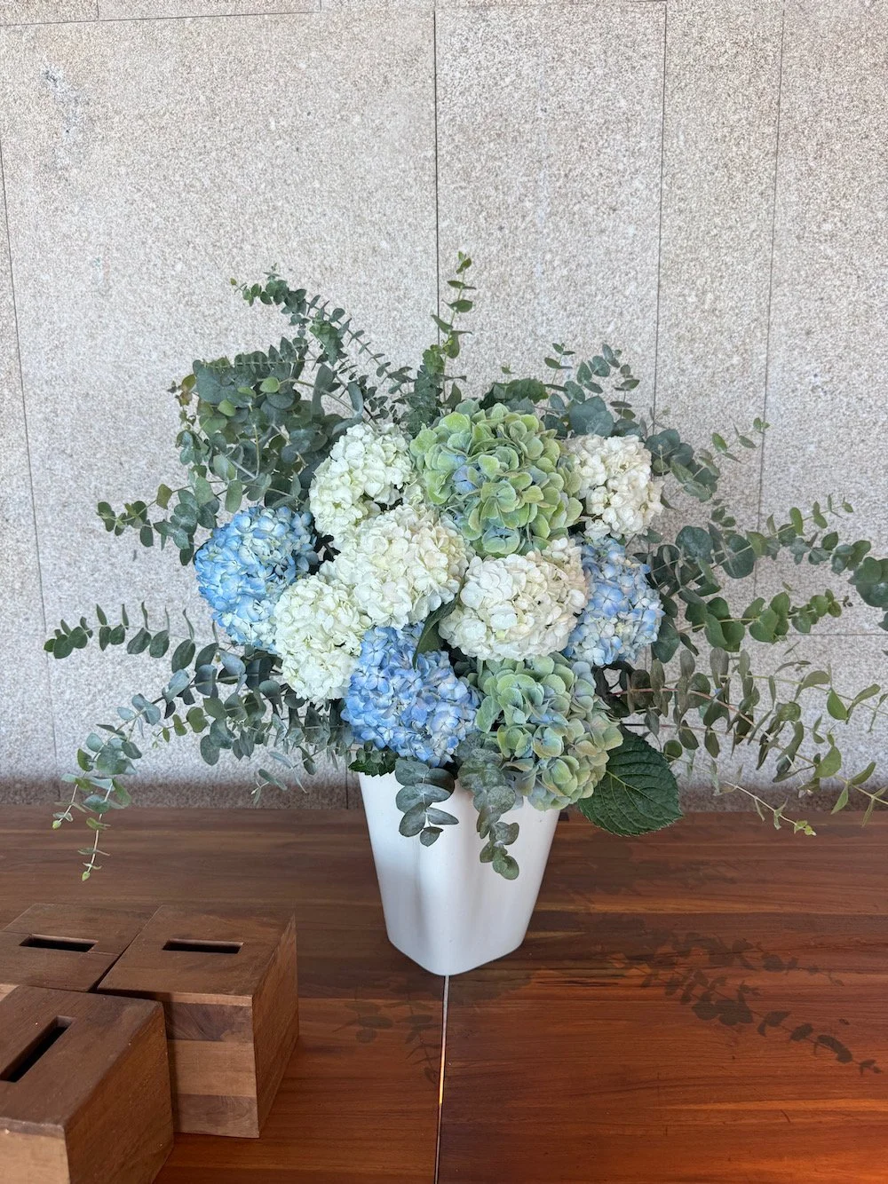 A bouquet of white, blue, and green hydrangeas with green foliage in a white vase on a wooden table.