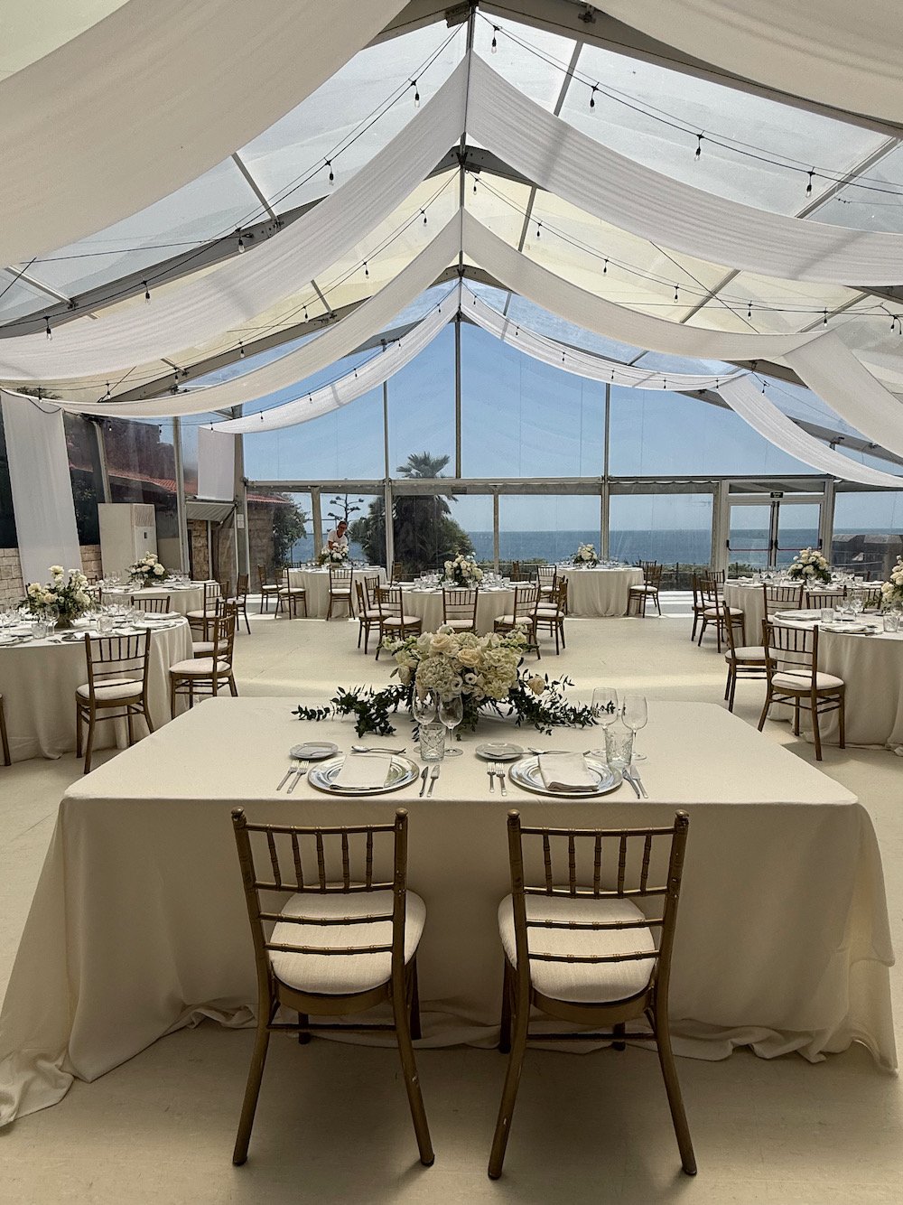 Elegant wedding reception tent with draped white fabric, string lights, and ocean view, featuring multiple round tables with white tablecloths, floral centerpieces, and place settings with plates, wine glasses, and silverware. In the center foregroun