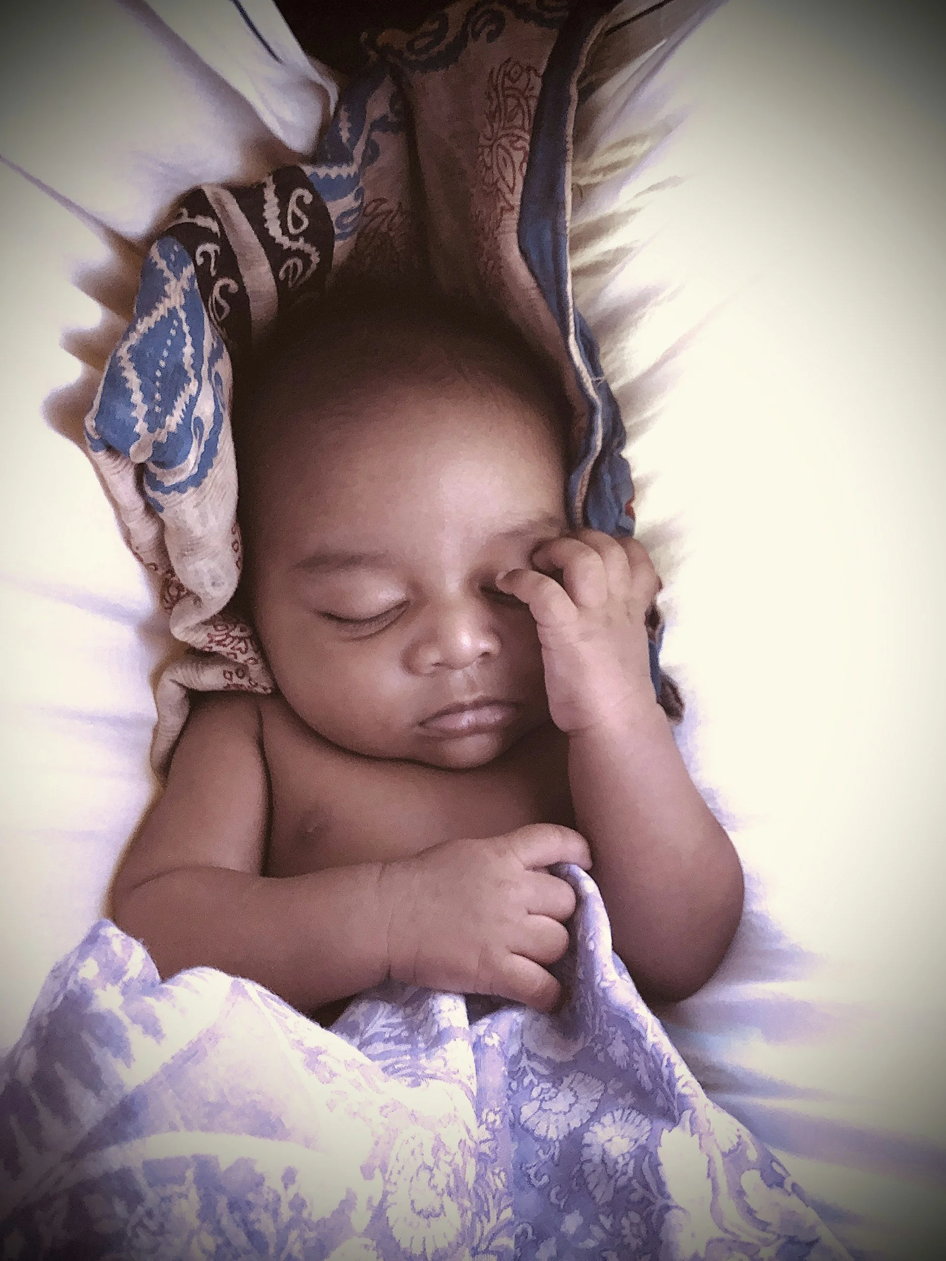 A sleeping baby lying in a bed, partially covered with a patterned blanket, with a colorful cloth head covering.