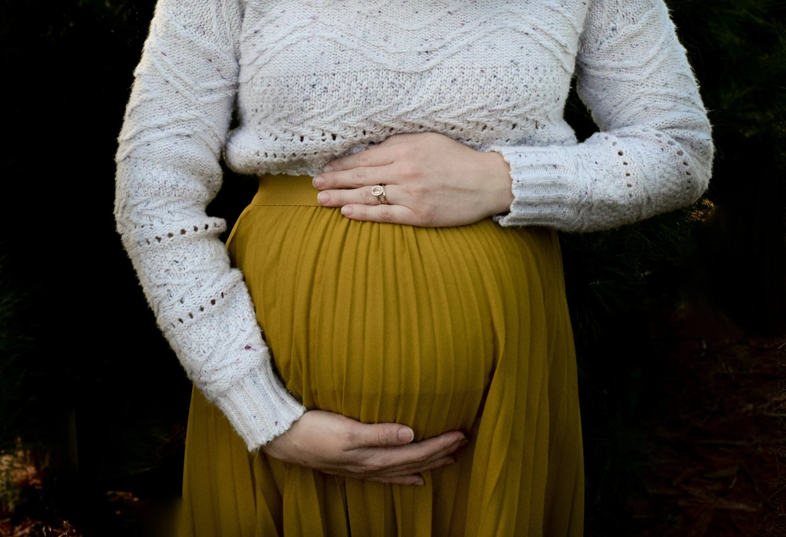 A pregnant woman wearing a white knitted sweater and a mustard-yellow pleated skirt, with her hands resting on her belly.