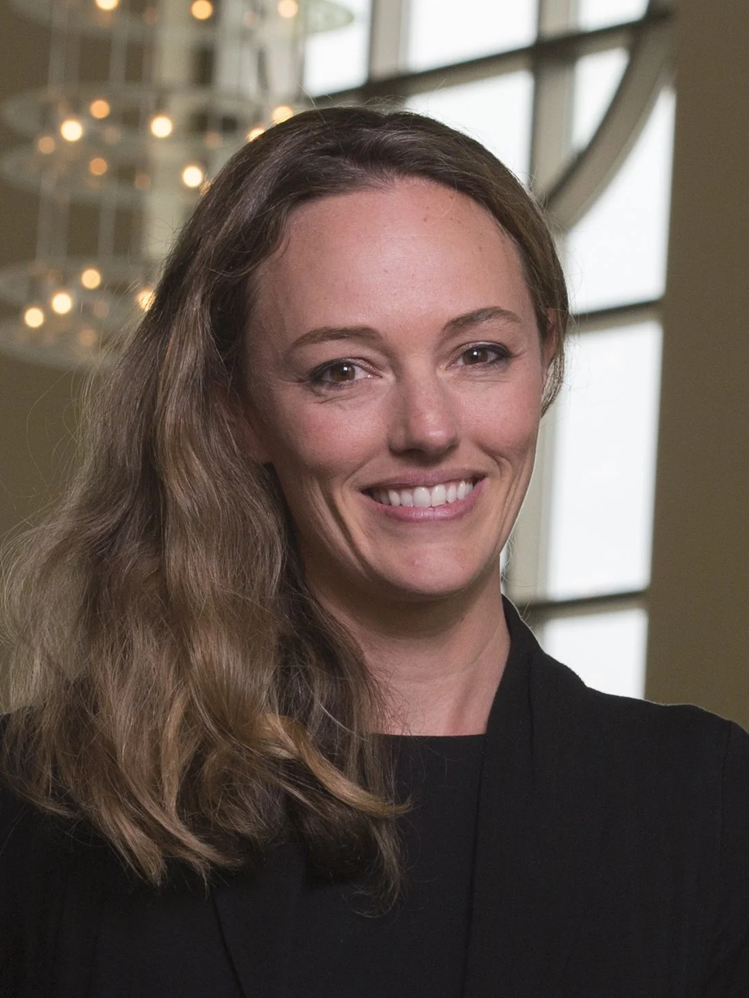 A woman with long wavy hair smiling indoors near large windows, with a chandelier in the background.