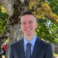 A young man in a suit smiling outdoors next to a large tree with green leaves.