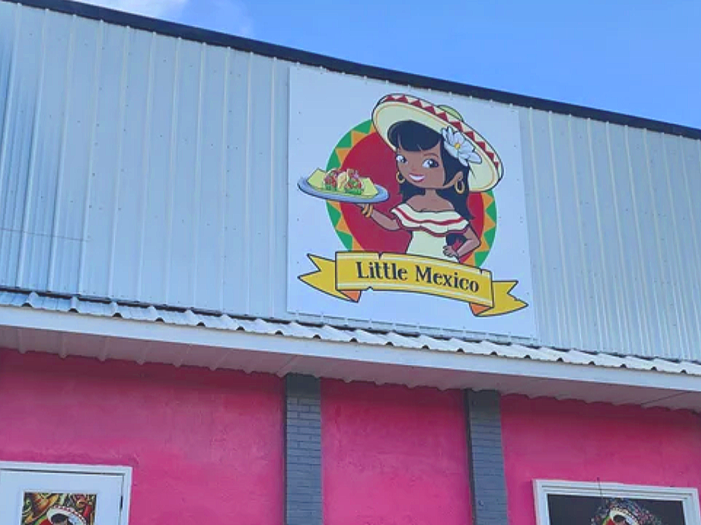 Sign for Little Mexico restaurant featuring a cartoon girl in a sombrero holding a plate of food, with a pink and blue building background.