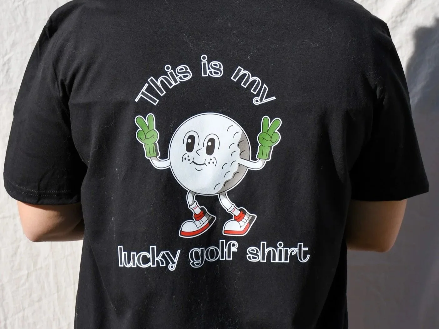 Back view of a person wearing a black T-shirt with a cartoon golf ball character making a peace sign with both hands. The shirt has white text that says "This is my lucky golf shirt."