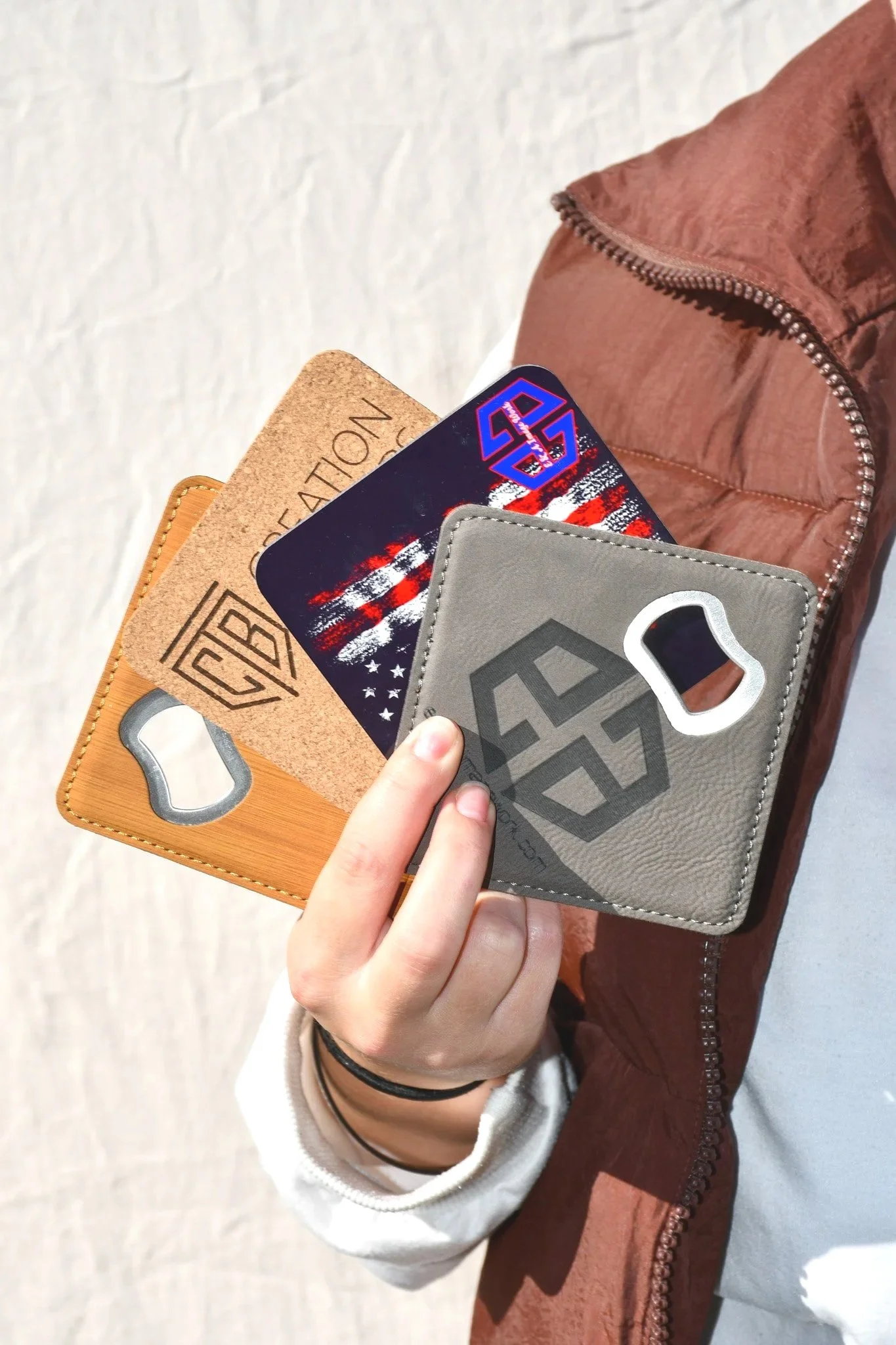 A person's hand holding four wallets and a beverage opener, with a brown jacket sleeve visible, against a white textured background.