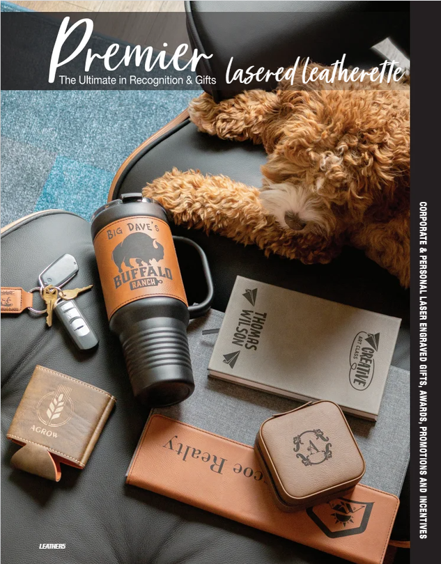 A puppy lying on a black leather surface next to a laser engraved metal cup with 'Big Dave's Buffalo Ranch' logo, a set of car keys, a wallet, a small leather box, a leather notebook, and a stack of books or brochures. The scene is part of a promotional display for laser engraved leather and gift products.