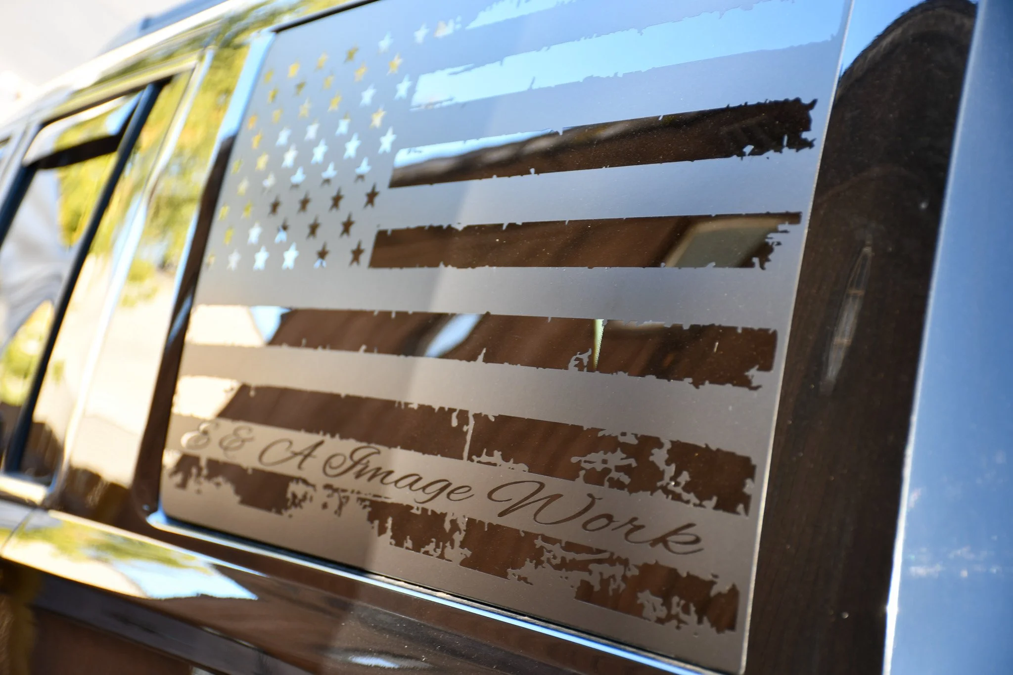 Car window sticker with an American flag and the words 'E & A Image N' work' written on it, with reflections of the surroundings visible.