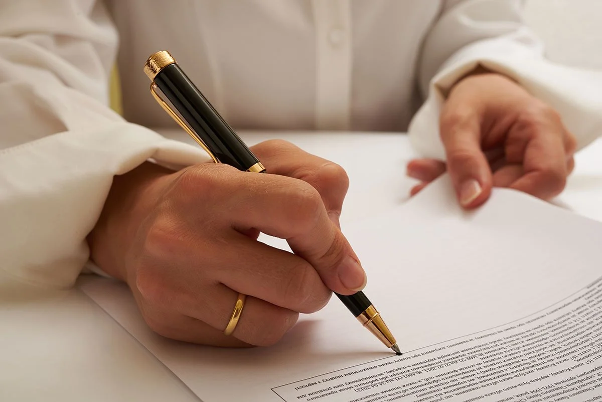 Close-up of a person signing a document with a black and gold pen, wearing a white shirt and a gold ring on their ring finger.