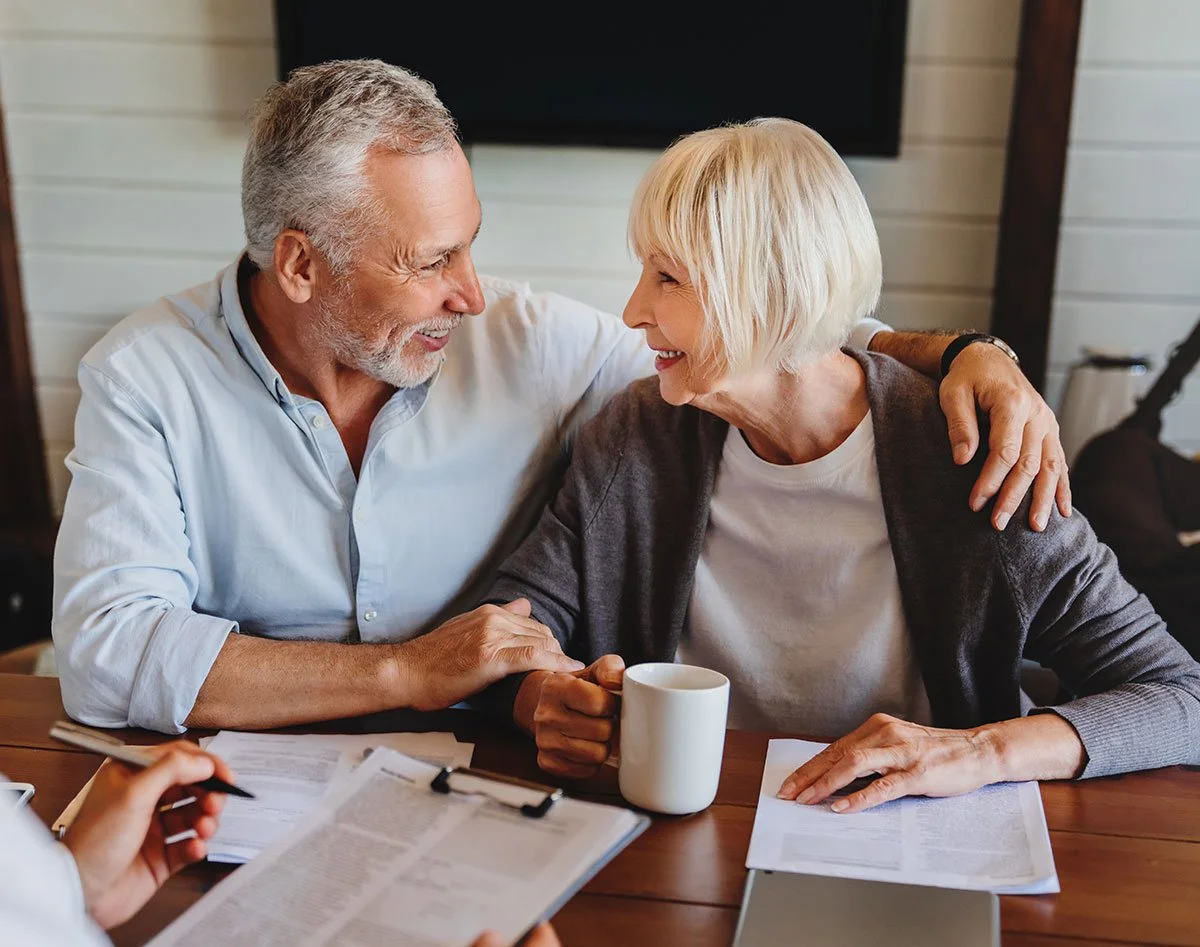 An elderly couple sitting at a table, smiling and looking at each other affectionately while holding hands, with documents and a coffee mug on the table.