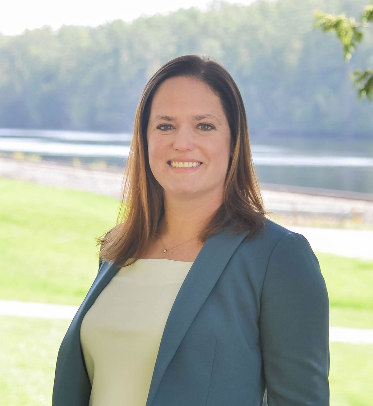 A woman with shoulder-length brown hair smiling outdoors with a river and trees in the background, wearing a dark blazer over a light-colored top.
