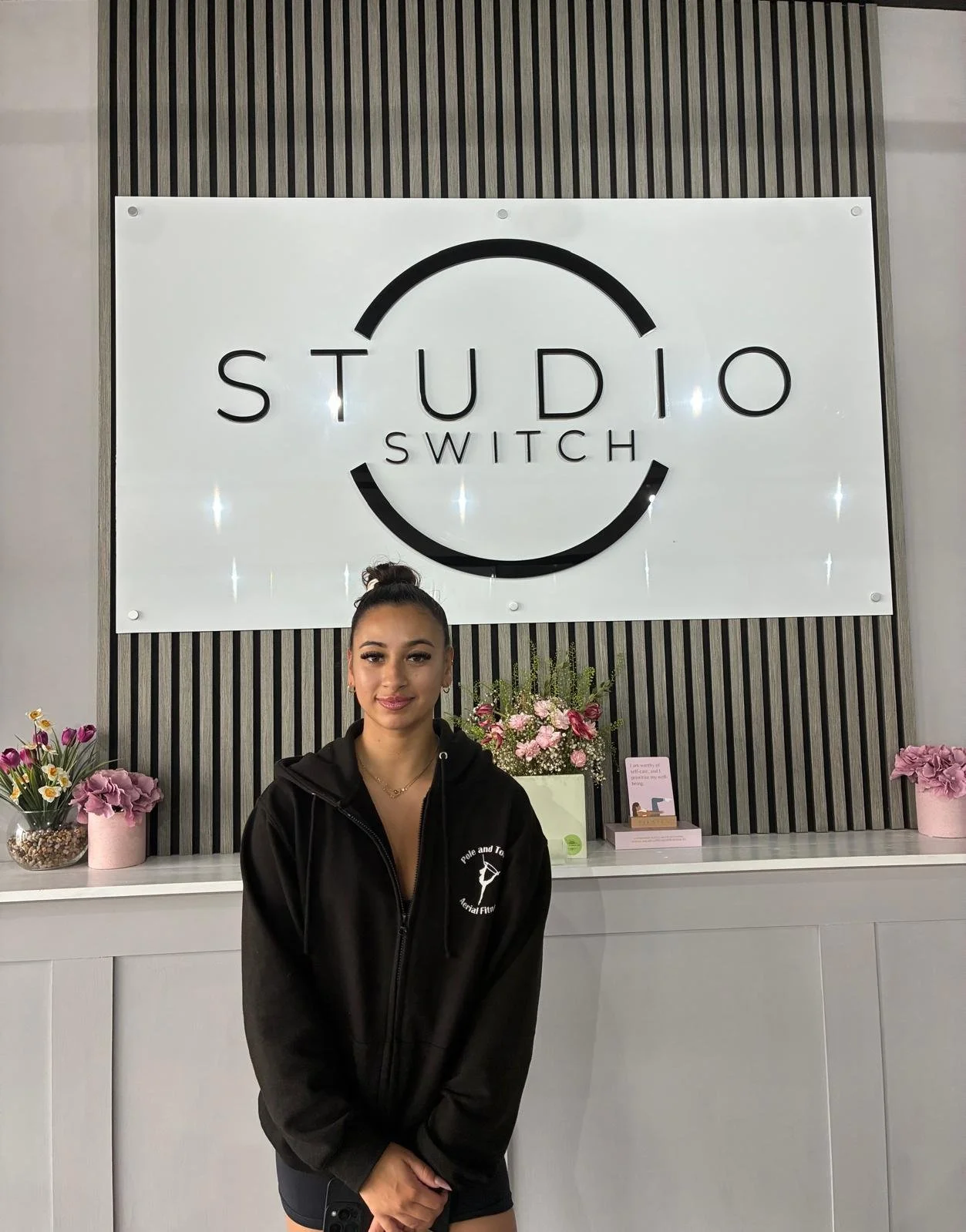 A young woman with dark hair in a bun, wearing a black zip-up hoodie with a logo, standing in front of a sign that reads 'Studio Switch' with floral decorations on a white counter.